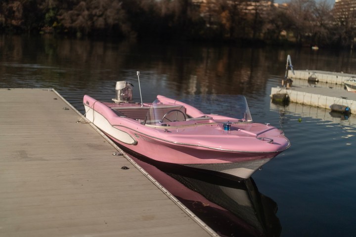A pink retro speedboat docked at a wooden pier on a calm lake.
