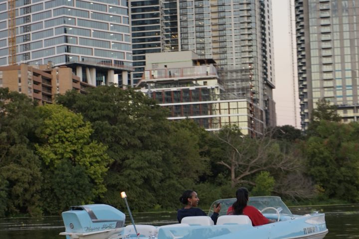 Two people in a blue boat on a river with tall city buildings in background.