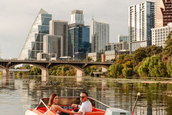 Couple on red boat on river with skyline and bridge in background.