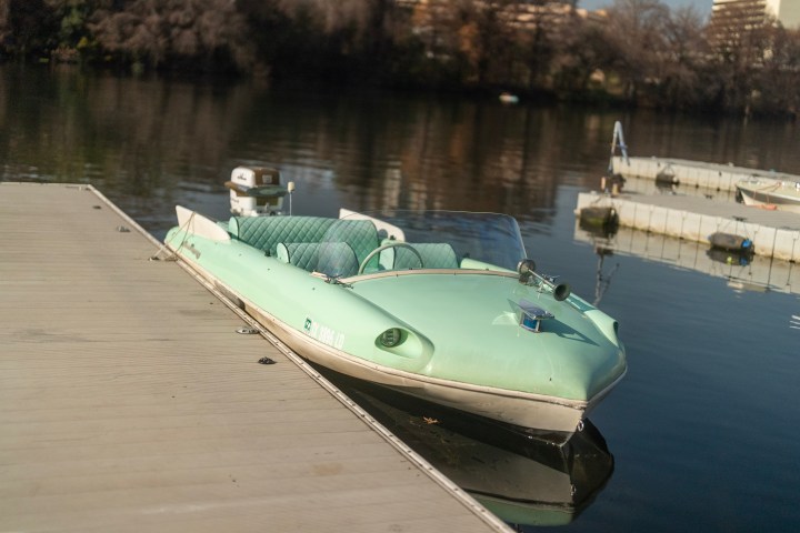 Seafoam green vintage boat docked on a calm lake with trees and buildings in the background.