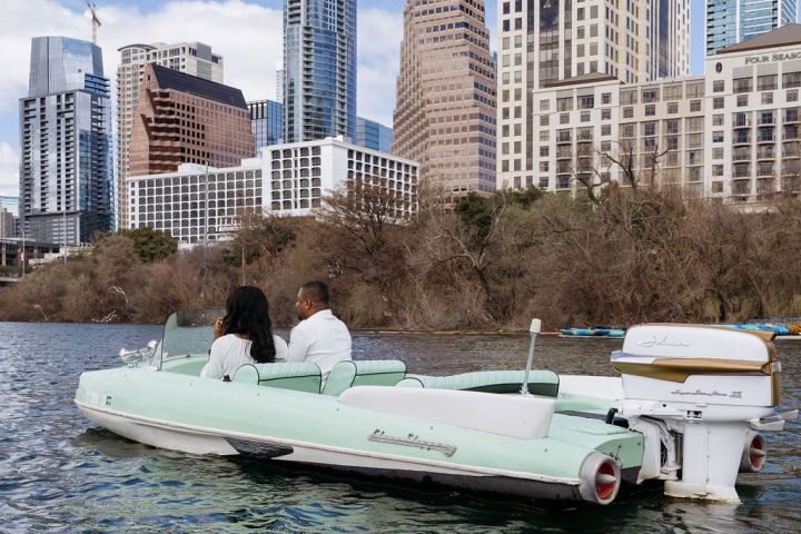 Two people sitting on a boat in a river with skyscrapers and a blue sky in the background.