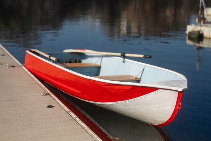 Red and white rowboat docked at waterside, with trees and a building in the background.