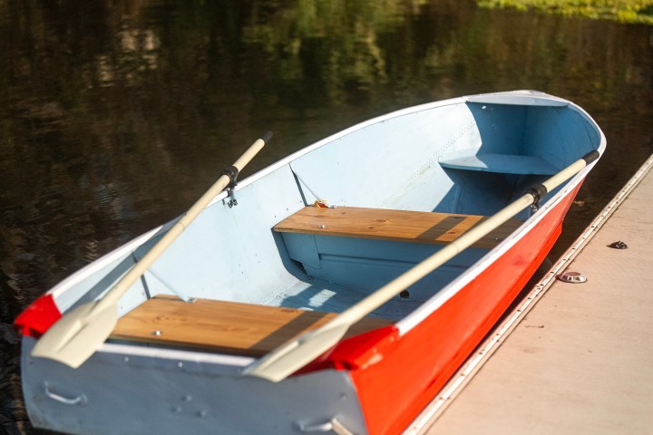 Small red and blue rowboat with oars tied to a dock on a calm lake.