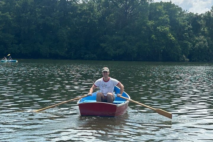 Person rowing a red boat on a lake with trees and clouds in the background.