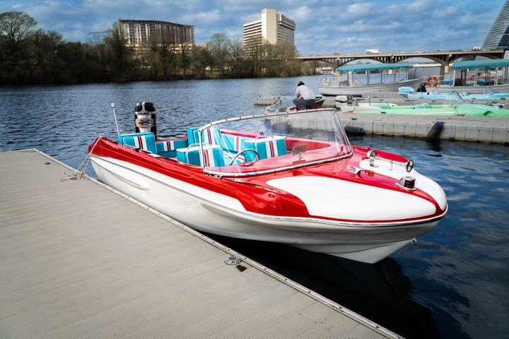 Red and white speedboat docked on calm water, with buildings in the background.